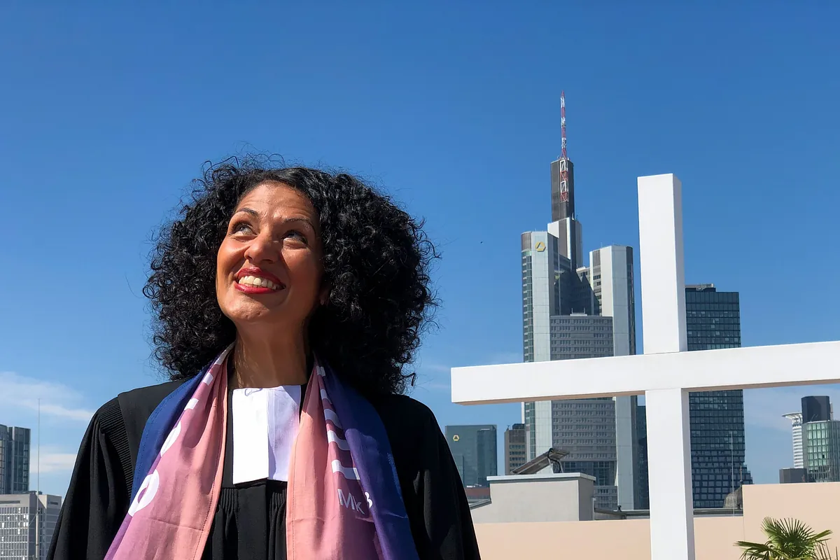 Sarah Vecera steht vor einem weißen Kreuz und der Frankfurter Skyline. Sie trägt ein buntes Halstuch und blickt gen Himmel.